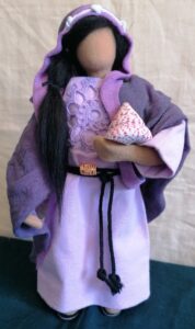 Handgefertigte Stoffpuppe in einem helllila Kleid mit violettem Umhang und Kapuze. Die Puppe hat langes schwarzes Haar und hält eine Purpurschnecke in ihrem Arm.